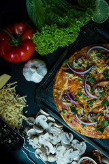 Fresh pizza with sausage, tomatoes, mushrooms and cheese on a dark background. Decorated with fresh vegetables and spices. View from above.