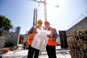 Structural engineer and construction manager in orange work vests and hard helmets discuss...