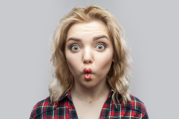 Portrait of funny amazed beautiful blonde young woman in casual red checkered shirt standing and looking at camera with big eyes and fish lips. indoor studio shot, isolated on grey background.