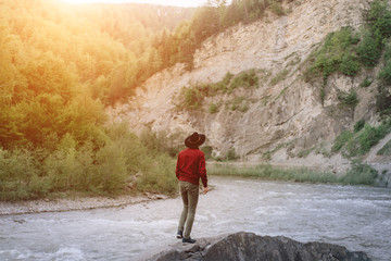 Obraz premium Young man in red jacket and black hat stands on a rock and enjoys mountain nature during the sunset. Concept outdoor nature lifestyle