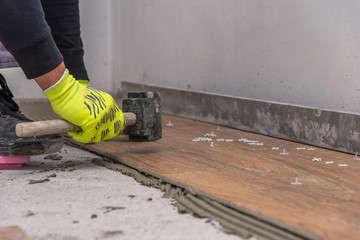 Worker placing ceramic floor tiles on adhesive surface, leveling with rubber hammer.