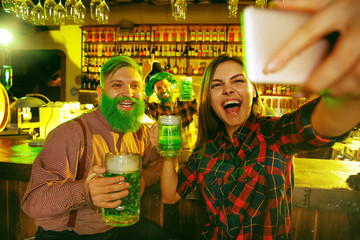 Saint Patrick's Day Party. Happy friends are celebrating and drinking green beer. Young men and women taking selfie photo with phone. Pub interior.