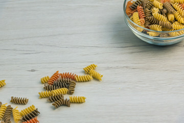 Raw uncooked three-colored Fusilli in a glas bowl on white wood table.