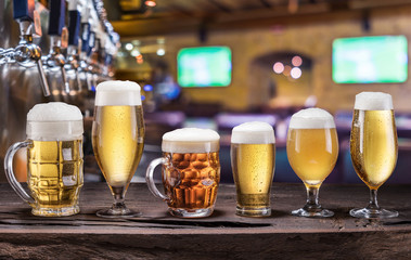 Chilled mugs and glasses of beer on the old wooden table. Pub interior and bar counter with beer taps at the background.