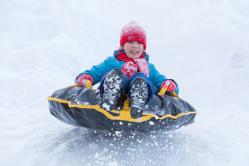 The girl in blue clothes with red hat and scarf descending from the hill covered with a snow on the black rubber ring instead of sledge. The winter picture of holiday recreation
