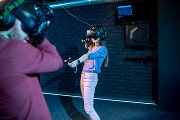 Man and woman playing game using virtual reality headset and gamepads in the dark room of the playing club