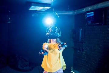 Young woman playing game using virtual reality headset and gamepads in the dark room of the playing club