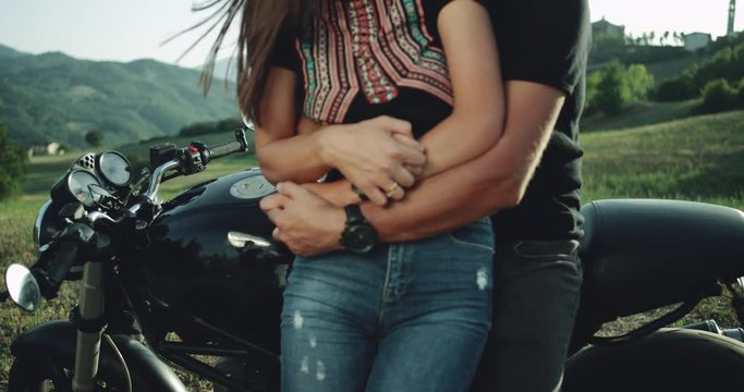Closeup details of a young couple in the middle of nature hugging each other behind of a black motorcycle in a sunny day