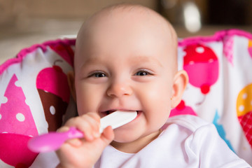 Baby in the kitchen in a baby chair holding a spoon and smiling.Sweet baby in high chair.Cheerful baby child eats food itself with spoon. Portrait of happy kid boy in high chair.first teeth 