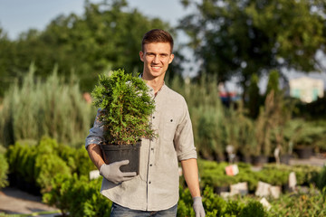 Guy gardener holds in his hand a pot with plant in the wonderful nursery-garden on a warm sunny day