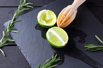 Lime halves and wooden juicer on slate board. Lime and rosemary on black background. Fresh lime...
