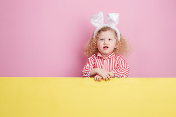 Funny little curly  girl in a striped red and white dress and bunny ears on her head stands  behind the yellow board against a pink wall