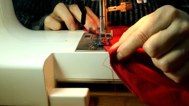 Process of tailoring working , hands of elderly seamstress sews clothes of red cloth with needle, looper and presser on a sewing machine. Small home business concept.