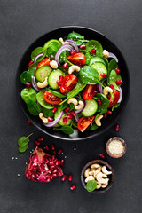 Spinach salad with fresh cucumbers, tomato, onion, pomegranate, sesame seeds and cashew nuts on black background. Healthy vegan food. Top view