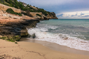 Wellen brechen sich weiß schäumend am sandig, felsigen Strand auf Mallorca