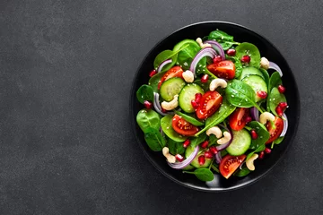 Poster Spinach salad with fresh cucumbers, tomato, onion, pomegranate, sesame seeds and cashew nuts on black background. Healthy vegan food. Top view © Sea Wave