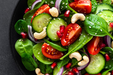 Spinach salad with fresh cucumbers, tomato, onion, pomegranate, sesame seeds and cashew nuts on black background. Healthy vegan food. Top view