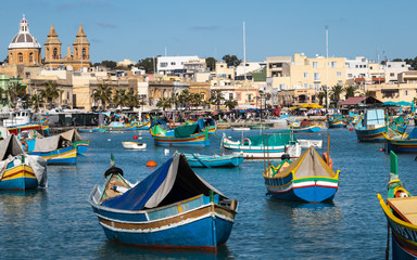 Fototapeta na wymiar Marsaxlokk Harbour with Traditional, colorful Luzzu Boats in the bay with market in background. Marsaxlokk, Malta, Europe.