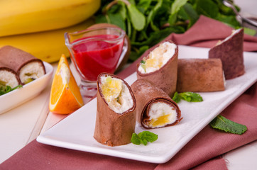 Sweet chocolate rolls from pancakes with cream cheese, fresh fruit and berry sauce on a white wooden background.
