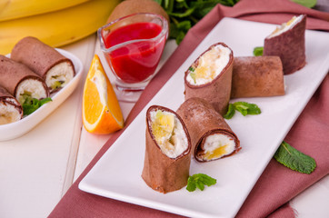 Sweet chocolate rolls from pancakes with cream cheese, fresh fruit and berry sauce on a white wooden background.