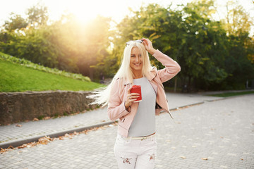 The concept of street fashion. young stylish girl student walking in the sunny park and holds a cup of coffee to go. portrait of smiling blond girl in sunglasses 