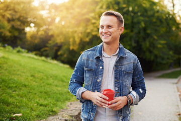 Young Hipster man standing with takeaway coffee in the park,  smiling plesantly into camera.  Happy...