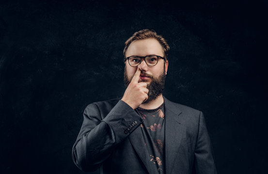 Portrait of a bearded guy in a formal jacket with a silly look picks a finger in his nose. Studio photo with dark wall background