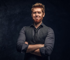 Smiling elegantly dressed redhead man posing with his arms crossed in studio against a dark textured wall