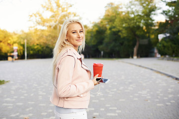 Sunny lifestyle fashion portrait of young stylish blond woman walking on street or park with phone, wearing trendy outfit, drinking coffee to go, smiling, enjoy weekends