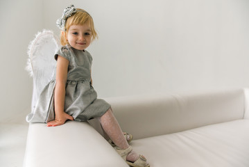 little cute little blonde girl in a gray dress with angelic white wings on a light sofa in the...