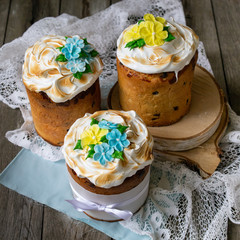 Three traditionally baked orthodox easter cakes with glace icing and bright blue and yellow flowers on woooden background with lace fabric. Easter kulich. Selective focus, copy space.