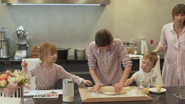 All the family is in the kitchen. The mother is busy at the back. The eldest daughter tries to roll the dough with the help of rolling pin. The other kids just mind their own business.