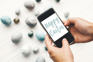Happy Easter text sign on phone screen in hands above stylish easter eggs on rustic white wooden table. Modern easter eggs painted with natural dye in grey marble. Easter greetings card
