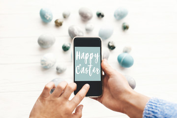 Happy Easter text sign on phone screen in hands above stylish easter eggs on rustic white wooden table. Easter greetings card online. Modern easter eggs painted with natural dye in grey
