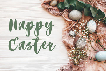 Happy Easter text sign on stylish easter eggs with spring flowers and eucalyptus on rustic fabric on white wooden table, flat lay. Easter greetings card