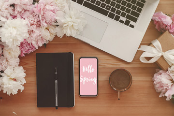 Hello Spring text sign on phone screen at black notebook, coffee, laptop, peonies on rustic wooden table flat lay. Freelance concept. Stylish floral greeting card.