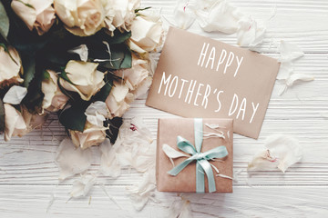 Happy Mother's Day text sign on craft greeting card and white roses bouquet, gift box on wooden background, flat lay. Mothers day. Floral greeting card