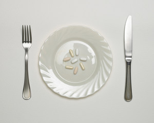 VITAMIN AND DIET SUPPLEMENT PILLS ON WHITE PLATE WITH KNIFE AND FORK