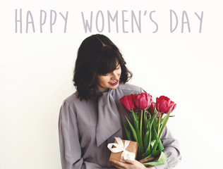 Happy Women's Day text sign and beautiful girl holding stylish bouquet of tulips and gift box on white background. Greeting card. International womens day 8 march