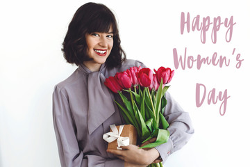 Happy Women's Day text sign and beautiful girl holding stylish bouquet of tulips and gift box on white background. Greeting card. International womens day 8 march