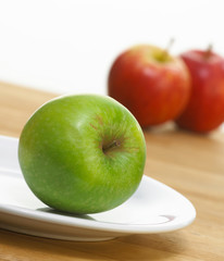 FRESH GREEN APPLE ON WHITE PLATE WITH RED APPLES IN BACKGROUND