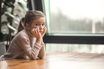 Charming little girl sitting at a wooden table by the window.