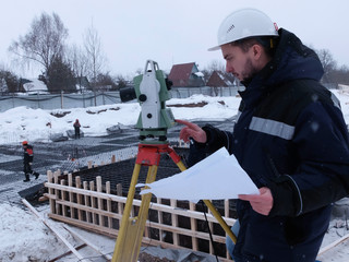 Bearded geodetic engineer-surveyor in white hardhat works on the geodetic total station holding a drawing sheet against the construction site at the background