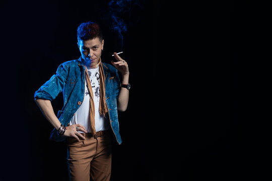A woman with red hair in a denim suit smokes a cigarette and releases a bym in blue light on a black background