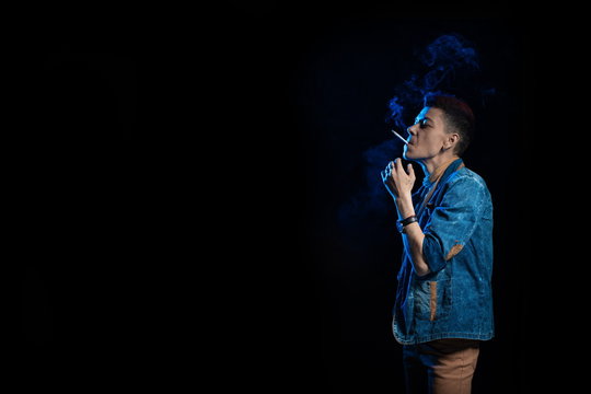 A woman with red hair in a denim suit smokes a cigarette and releases a bym in blue light on a black background