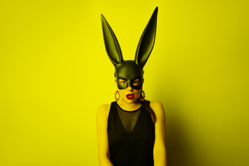 Sexy young woman with red lipstick and large breasts wearing a black mask. Easter bunny standing on a yellow background and looks very sensually.