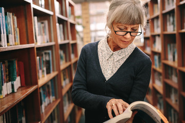 Senior woman looking at a book standing in library - obrazy, fototapety, plakaty
