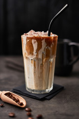 Ice coffee with cream in a tall glass and coffee beans on dark concrete table over black wooden background. Cold summer drink. Copy space for text. Selective focus.