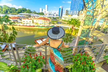 Tourist woman with wide hat looking panorama from terrace over Clarke Quay and Riverside area in Singapore, Southeast Asia.Cruise on Singapore River in sunny day and skyscrapers downtown on background - obrazy, fototapety, plakaty