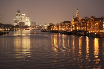 Fototapeta na wymiar AMSTERDAM, NETHERLANDS - NOVEMBER 21, 2018: Illuminated buildings reflected in calm water, amazing cityscape with boat on Amstel canal at night